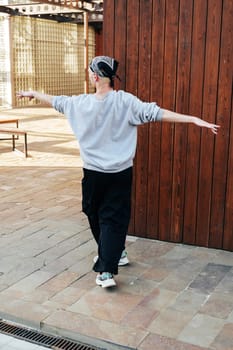 A person walks away from a wooden wall. They are wearing a grey sweatshirt, black pants, and a black bandana. Their arms are stretched out to the sides.