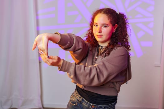 A young woman with curly brown hair is posing in a studio. She is wearing a brown sweater, a black top, and blue jeans. She has one arm extended, and her hands are forming a frame.