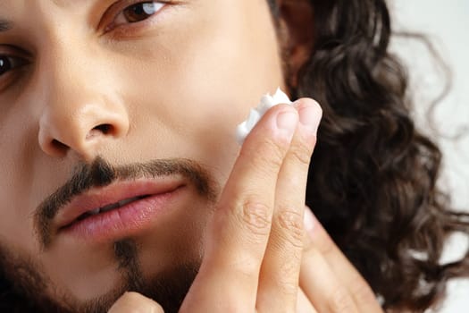 A young man applying skincare cream on his face with a focused expression, demonstrating personal grooming at home close up