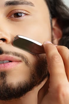 Man with curly hair grooming his beard carefully using a straight razor against a plain white background close up