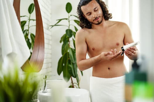A young man applies skincare cream while looking in a round mirror in a bright bathroom close up