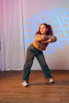 A young woman with curly red hair is dancing in a studio. She is wearing blue jeans and a light brown jacket. Her arms are crossed and she is looking to the side.