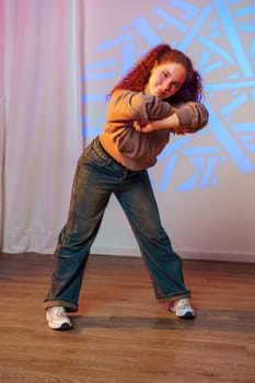 A young woman with red hair and a brown jacket dances in a studio, her body positioned in a dynamic pose, one leg extended forward and the other bent at the knee.