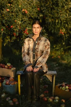 Stylish girl in black clothes sitting near picked apples in a sunny fall orchard.