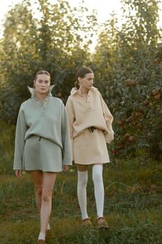 Two models in stylish tracksuits pose in apple orchard at sunset . The model is wearing a light-colored tracksuit and skirt.