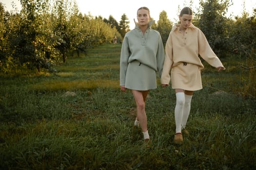 Two models in stylish tracksuits pose in apple orchard at sunset . The model is wearing a light-colored tracksuit and skirt.