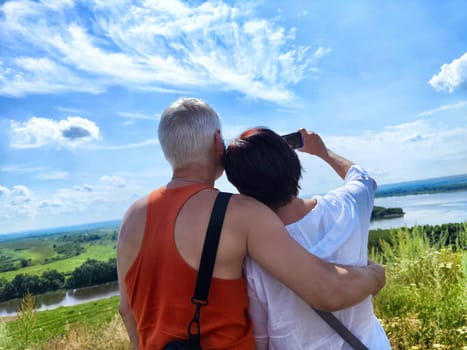 A couple stands together on a hill, embracing and taking a selfie with a picturesque landscape and river in the background.
