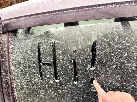 A person writes a friendly greeting in snow covering a car window on a cold winter day in a suburban neighborhood.