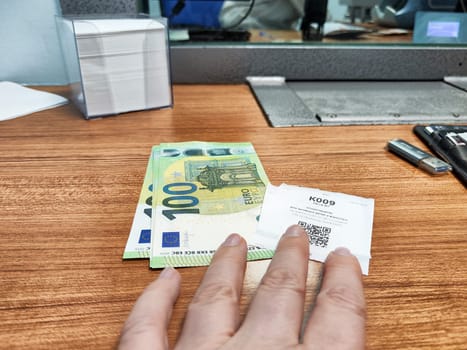 Kirov, Russia - October 08, 2024: A hand reaching for a currency voucher next to euro banknotes on a wooden counter at a currency exchange location