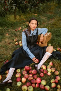 Portrait of a beautiful Asian woman in stylish clothes with apples in the garden. The girl is sitting on the ground and there are a lot of red apples around.