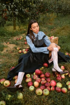 Portrait of a beautiful Asian woman in stylish clothes with apples in the garden. The girl is sitting on the ground and there are a lot of red apples around.