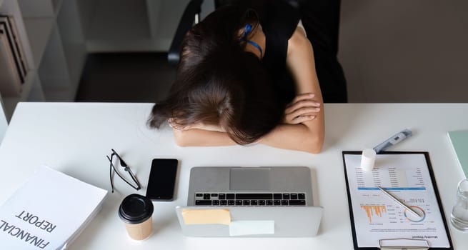 A young businesswoman appears tired and stressed, resting her head on her arms at a cluttered office desk.
