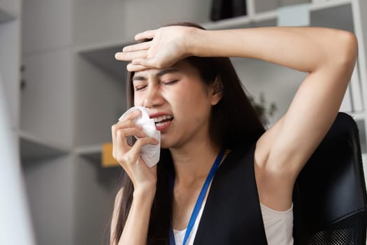 A businesswoman appears sick at her office desk, holding a tissue and looking distressed, highlighting workplace health challenges.