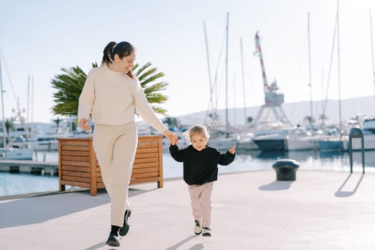 Smiling mother and little girl are skipping along the pier holding hands. High quality photo
