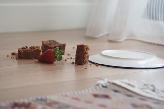 An image depicting a fallen plate on a floor with chocolate dessert pieces and a strawberry.