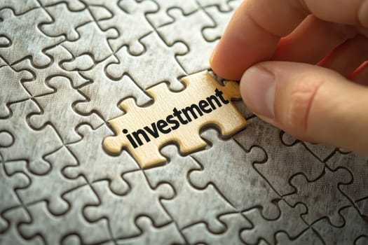 A close-up image of a hand placing a wooden puzzle piece labeled investment into a larger puzzle. This symbolizes the importance of financial planning and strategy.