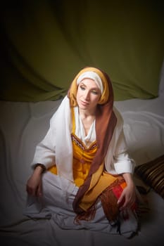 The girl, wearing vibrant national attire, sits gracefully, embodying Rebekah from the Christian Bible in a serene indoor setting.