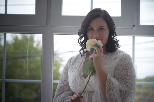 A plump woman holds a rose, dressed in a delicate gown, smiling softly in a sunlit room filled with natural light.