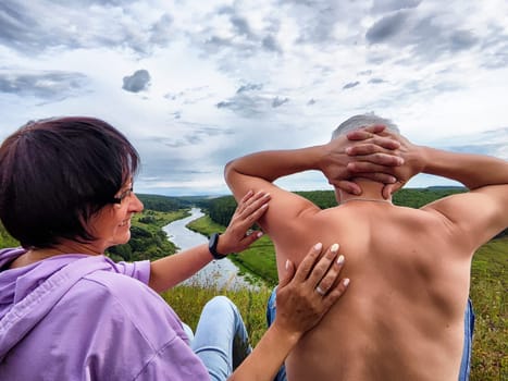 A woman massages a man's shoulders as they relax overlooking a river surrounded by lush greenery under a cloudy sky.