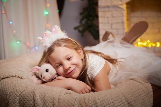 A young girl relaxes on a cozy surface, smiling with her plush toy in a warmly lit, decorated room celebrating the holidays.