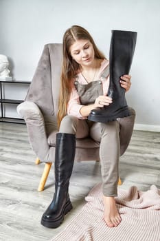 A young woman tries on a pair of trendy black knee-high boots while sitting comfortably in a modern chair at her home.
