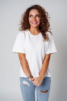 Cheerful woman with curly hair wearing a white shirt and jeans poses happily against a light gray background close up