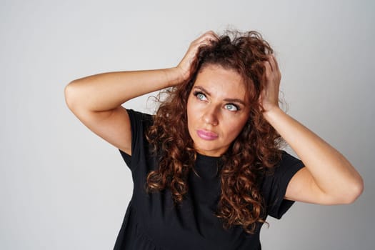 Smiling woman with curly hair poses confidently against a plain background while holding her hands behind her head indoors