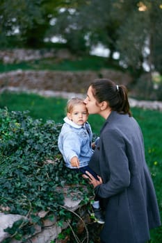 Mom kisses the temple of a little girl sitting on a fence in the park. High quality photo