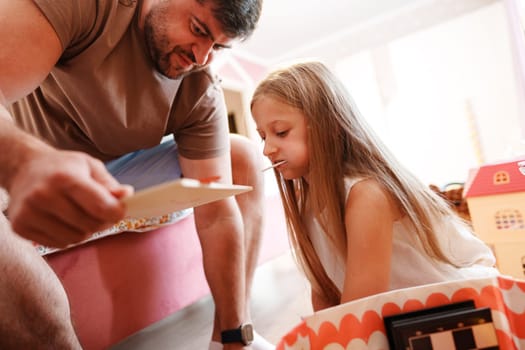 A man sits on a bed, leaning down to look at a board game. His daughter sits beside him, her face focused on the game. A toy house can be seen in the background.