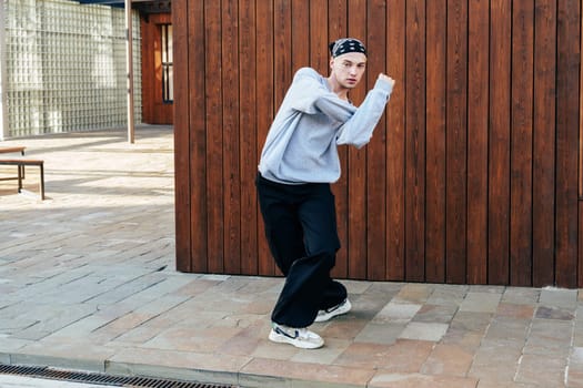 A young man in a grey sweatshirt and black pants, wearing a black bandana on his head, dances in front of a brown wooden wall.