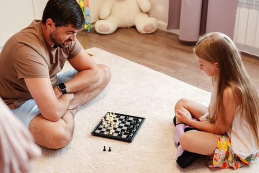 A man and young girl sit on a white rug in a living room. They are playing chess, and the man is looking at the chessboard while the girl sits with her legs crossed.