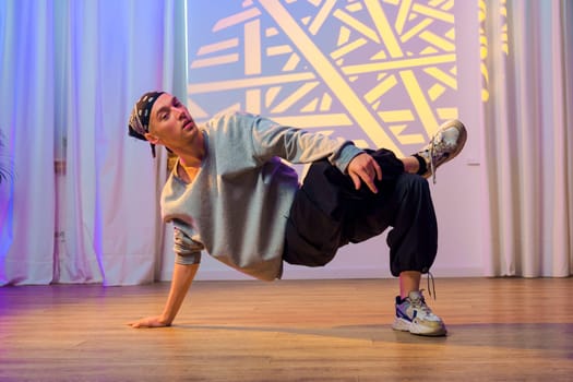 A dancer in a gray sweatshirt and black pants performs a breakdancing move on a wooden floor. Behind the dancer are white curtains and a light projection with a geometric pattern on the wall.