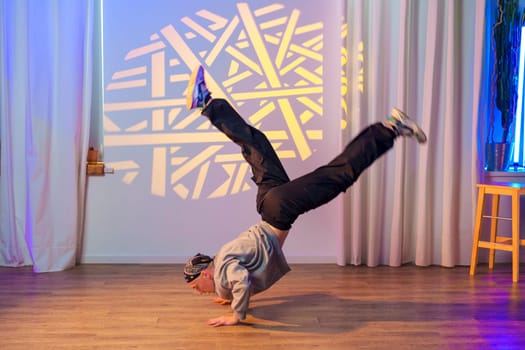 A young man wearing a grey sweatshirt and black pants performs a dance move in a studio. He is standing on his hands with his legs extended in the air.