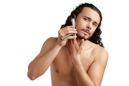 A man with curly hair grooming his beard in a well-lit space while using a straight razor for a clean shave