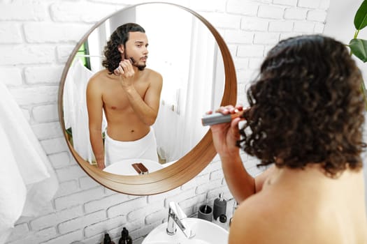 A man with curly hair grooming himself in the bathroom while looking into a round mirror close up