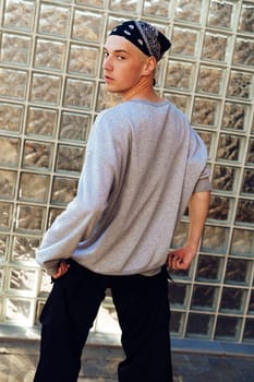 A young man stands in front of a brick wall, looking over his shoulder. He wears a gray sweatshirt, black pants, and a bandana tied around his head.