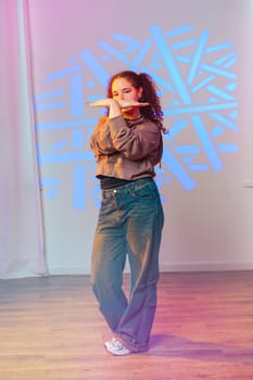 A young woman with curly hair is wearing a brown hoodie and blue jeans. She is standing with her arms crossed in front of her, looking directly at the camera.