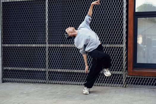 A young dancer, wearing a grey sweater and black pants, executes a dynamic dance move in front of a chain-link fence.