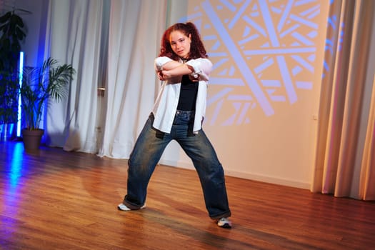 A woman with curly red hair and a white shirt dances in a studio. She has a casual look with her blue jeans and white sneakers.