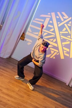 A young person dances in a studio with white curtains and wood flooring. The dancer wears a grey sweater and black pants, and a patterned light shines on the wall behind them.
