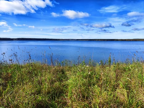 A serene lake stretches out beneath a blue sky with scattered clouds, framed by a grassy hillside.