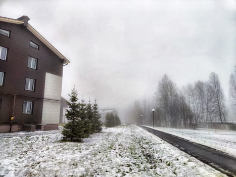 Snow blankets the ground as a dense fog envelops the pathway beside a residential building, creating a serene winter atmosphere.