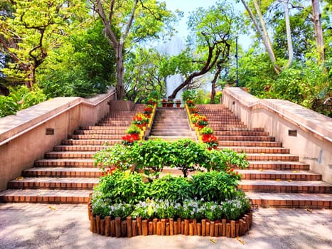 A lush garden features colorful flowers and greenery along stone steps leading to a tranquil fountain in a city park on a sunny day.