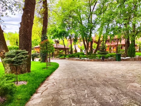 A beautiful stone pathway meanders through vibrant greenery and trees, leading to charming buildings in a peaceful garden.