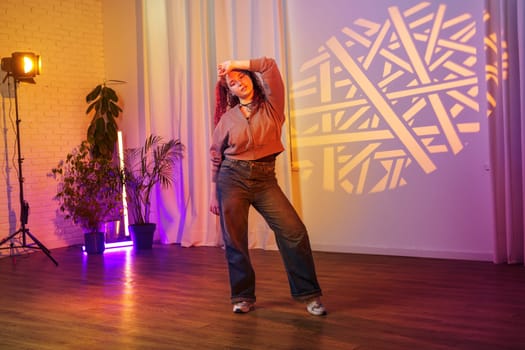 A young woman with long, dark hair dances in a studio. She is wearing a brown jacket and blue jeans, with her hand on her head.