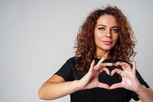 A young woman with curly hair makes a heart shape with her hands against a neutral backdrop close up