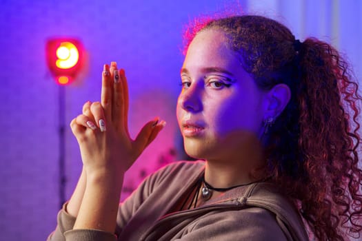 A young woman with long, curly hair, dressed in a brown hoodie and a black choker necklace, poses in a studio with colorful lights.