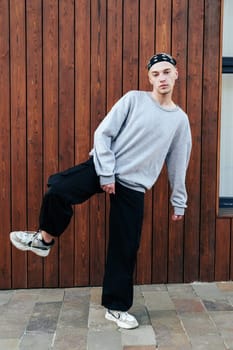 A young man wearing a gray sweatshirt, black pants, and white sneakers is standing in front of a wooden wall. He has a bandana on his head and his right leg is raised.