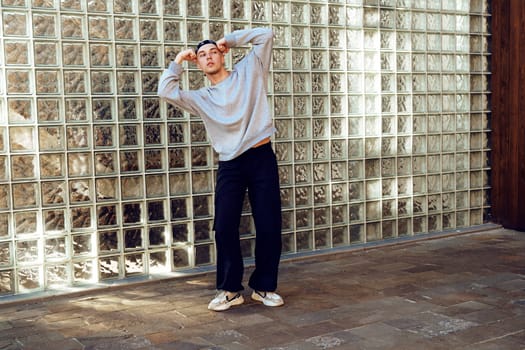 A young man, wearing a gray sweater and black pants, stands with his back to a glass block wall. The sunlight shines through the glass, creating a bright pattern on the ground.