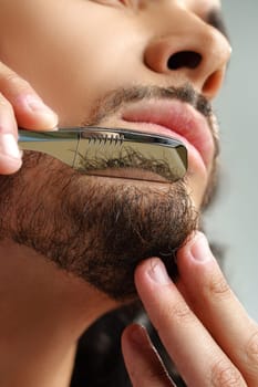 Man with curly hair grooming his beard carefully using a straight razor against a plain white background close up